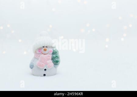 Dekorative Figuren im Weihnachtsstil. Die Statuette eines Schneemanns in einem gestrickten Hut auf weißem Hintergrund. Festliche Einrichtung, warme Bokeh-Lichter Stockfoto