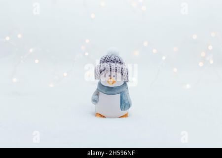 Dekorative Figuren im Weihnachtsstil. Die Statue eines Pinguins in einem gestrickten Hut auf weißem Hintergrund. Festliche Einrichtung, warme Bokeh-Lichter Stockfoto