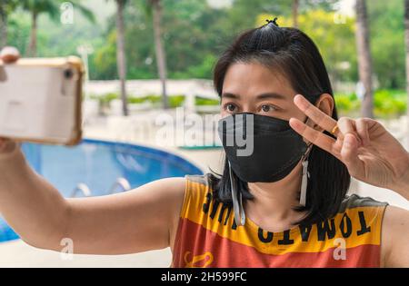 Portrait glückliche asiatische Frau mittleren Alters mit schwarzer Gesichtsmaske macht Selfie in einem Resort während der Reise in Thailand, zeigt zwei Finger Geste Stockfoto