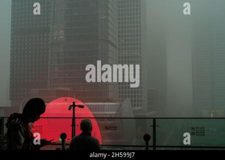 Ein junger Mann überprüft sein Mobiltelefon, während ein anderer den Wolkenkratzer, der in starkem Smog gehüllt ist, im chinesischen Geschäftsviertel von Peking ansieht. 06-Nov-2021 Stockfoto