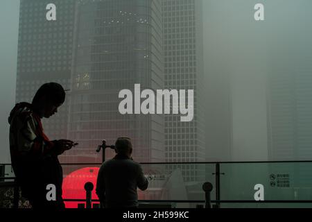 Ein junger Mann überprüft sein Mobiltelefon, während ein anderer den Wolkenkratzer, der in starkem Smog gehüllt ist, im chinesischen Geschäftsviertel von Peking ansieht. 06-Nov-2021 Stockfoto