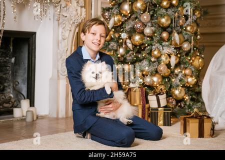 Hübscher kleiner blonder Junge mit jungen Haaren, fröhlich lächelndes Gesicht, in blauem Anzug gekleidet. Spielt mit einem pommerschen Welpen. Porträt eines Kindes und Tiere. Weihnachtsurlaub für die ganze Familie. Stockfoto