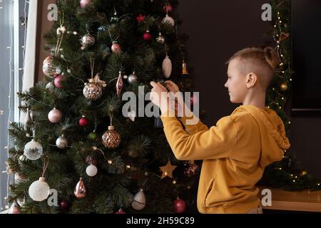 Junge in gelber Kapuzenjacke schmückt Baum im Wohnzimmer während der Vorbereitung auf Weihnachten Stockfoto