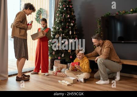 Kinder öffnen Geschenkboxen an geschmückten Tannenbäumen und genießen Weihnachtsgeschenke, die von den Eltern geschenkt werden Stockfoto