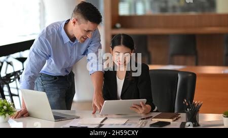 Glückliche asiatische Geschäftsfrau reden und arbeiten auf Tablet mit kaukasischen männlichen Chef im modernen Büro. Stockfoto