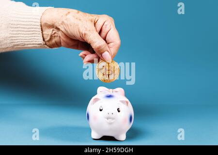 Faltige Hand einer älteren Frau legt eine Bitcoin in ein Sparschwein. Blauer Hintergrund. Das Konzept der Investition und Kryptowährung. Stockfoto