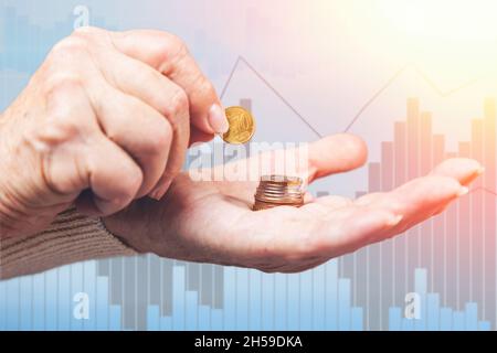 Nahaufnahme von Frauen mit faltigen Händen, die eine Euro-Münze in einen Münzstapel gelegt haben. Business Chart im Hintergrund. Das Konzept von Armut und Ersparnissen. Stockfoto
