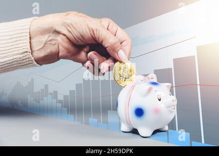 Faltige Weibchen legen eine Bitcoin ein Sparschwein. Business Chart im Hintergrund. Sparschwein ist entkokkkkkkkkk“. Das Konzept der Investition und des Sparens. Stockfoto