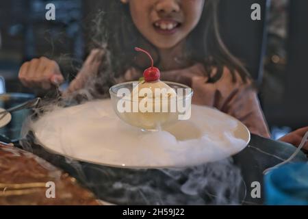 Ein süßes, junges asiatisches Mädchen sitzt in einem Restaurant und isst eine Schüssel Vanilleeis, dekoriert mit weißem Rauch aus Trockeneis, mit Kirschenauflage. K Stockfoto