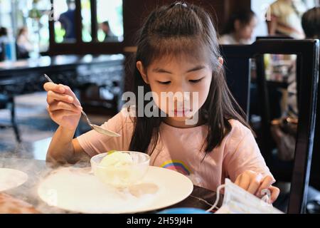 Ein süßes, junges asiatisches Mädchen sitzt in einem Restaurant und isst eine Schüssel Vanilleeis, dekoriert mit weißem Rauch aus Trockeneis, mit Kirschenauflage. K Stockfoto