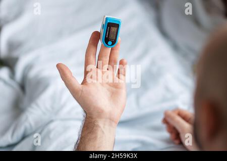 Patient liegt am Bett und überprüft die Daten am Pulsoximeter. Draufsicht. Konzept der Bestimmung von Sauerstoff im Blut. Stockfoto