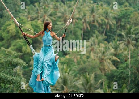 Gebräunte schöne Frau in einem langen türkisfarbenen Kleid mit einem Zug, die auf einer Schaukel reitet. Im Hintergrund ein Regenwald und Palmen. Speicherplatz kopieren. Stockfoto