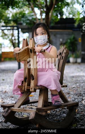 Ein süßes junges asiatisches Mädchen mit einer Gesichtsmaske über ihrem Mund reitet auf einem pferdeförmigen hölzernen Schaukelstuhl auf einem Spielplatz während Covid-19 pand Stockfoto