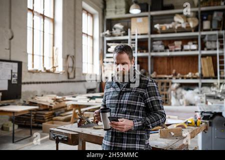 Porträt eines reifen männlichen Zimmermanns mit Kaffee und Smrtphone in Innenräumen in der Schreinerei. Stockfoto