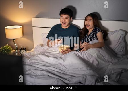 Lustiges junges Paar, das Fernsehen schaut und nachts auf einem Bett lacht Stockfoto