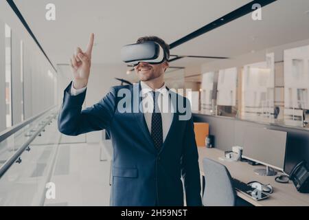 Glücklicher Büroangestellter, der eine vr-Brille trägt und mit dem Finger auf Objekte in der digitalen Welt zeigt Stockfoto