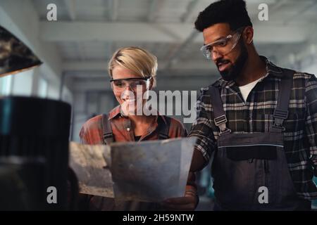 Porträt junger birazialer Industriekollegen, die in Innenräumen in einer Metallwerkstatt arbeiten, lächelnd. Stockfoto