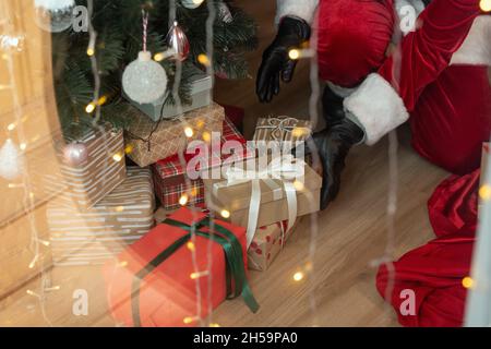 Nahaufnahme des nicht erkennbaren Weihnachtsmannes in schwarzen Handschuhen, der Weihnachtsgeschenke unter den Baum legt, Weihnachtslichteffekt Stockfoto