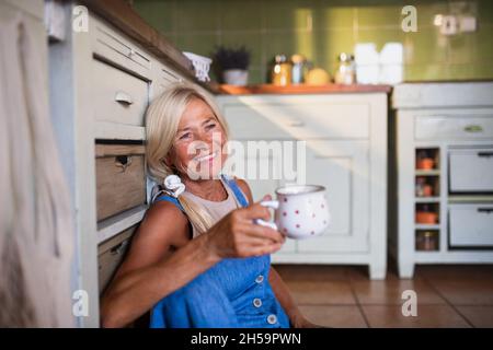 Glückliche ältere Frau, die auf dem Boden sitzt und in der Küche Tee trinkt. Stockfoto