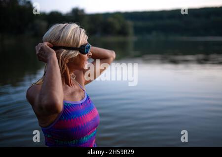 Porträt einer aktiven älteren Schwimmerin, die im Freien im See steht und eine Brille aufsetzt. Stockfoto