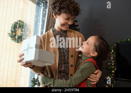 Happy hübsches Mädchen umarmt Mutter mit Geschenkbox, während sie zu Weihnachten gratuliert Stockfoto