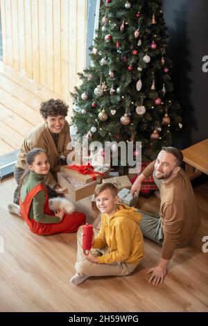 Porträt der positiven schönen kaukasischen Familie sitzt mit Geschenken auf dem Boden gegen Weihnachtsbaum zu Hause Stockfoto