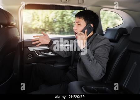 Gestresster junger Geschäftsmann, der auf dem Rücksitz des Autos sitzt und ein Problem mit einem Mobiltelefon spricht Stockfoto