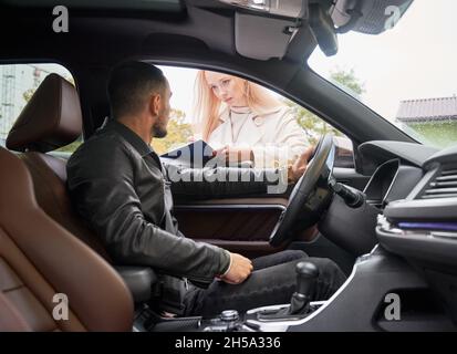 Junger, gutaussehender, glücklicher Mann, der in einem neuen Auto sitzt. Schöne Verkäuferin, die darum bittet, im Autohaus im Freien einige wichtige Dokumente zu unterschreiben. Blick aus dem Inneren eines Autos. Stockfoto