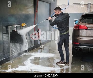 Self-Service-Waschstation. Junger männlicher Fahrer, der hinter seinem Auto steht und in den Händen Spezialausrüstung zum Waschen des Fahrzeugs hält. Mann mit starken Wasserstrahl Reinigung Auto Matten. Stockfoto