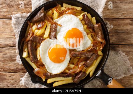 Chorrillana ist ein Gericht, das aus pommes frites mit geschnittenem Fleisch, Spiegeleiern und Zwiebeln in der Pfanne auf dem Tisch besteht. Horizontale Ansicht von oben Stockfoto