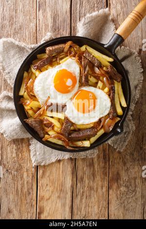 Chorrillana ist ein beliebtes chilenisches Gericht mit Pommes frites mit Rindfleisch, karamellisierten Zwiebeln und Spiegeleiern aus der Nähe in der Pfanne auf dem Tisch. Vertikal oben vi Stockfoto