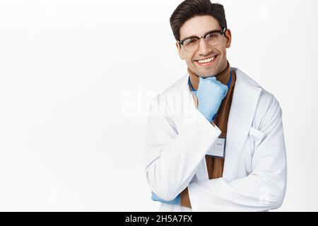 Lächelnder Arzt in Brille und medizinischen Handschuhen, der den Patienten ansieht, fröhlicher Gesichtsausdruck, medizinische Kleidung, weißer Hintergrund Stockfoto