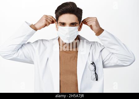 Covid 19. Hübscher Brünette Arzt setzt auf medizinische Gesichtsmaske vor der Untersuchung des Patienten, stehend über weißem Hintergrund Stockfoto