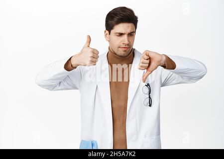 Unentschlossene Ärztin, die den Daumen nach oben und unten zeigt, kann sich nicht entscheiden, sie entscheidet sich und steht in weißer Robe vor weißem Hintergrund Stockfoto