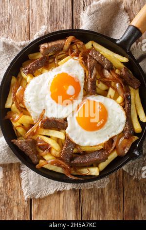 Chilenische Chorrillana Pommes mit Rindfleisch, karamellisierten Zwiebeln und Spiegeleiern aus der Nähe in der Pfanne auf dem Tisch. Vertikale Draufsicht von oben Stockfoto