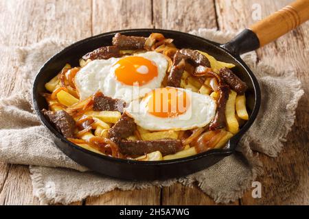 Chorrillana ist ein beliebtes chilenisches Gericht mit Pommes frites, die mit Rindfleisch, karamellisierten Zwiebeln und Spiegeleiern aus der Nähe in der Pfanne auf dem Tisch gefüllt sind. Horizont Stockfoto