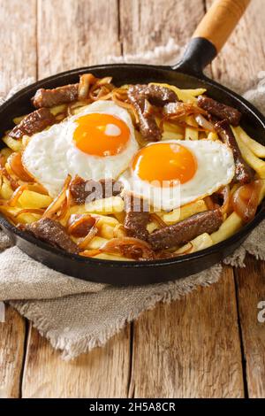 Chilenische Chorrillana Pommes mit Rindfleisch, karamellisierten Zwiebeln und Spiegeleiern aus der Nähe in der Pfanne auf dem Tisch. Vertikal Stockfoto