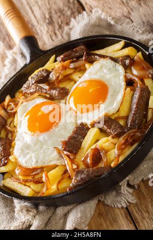 Chorrillana ist ein beliebtes chilenisches Gericht mit Pommes frites mit Rindfleisch, karamellisierten Zwiebeln und Spiegeleiern aus der Nähe in der Pfanne auf dem Tisch. Vertikal Stockfoto