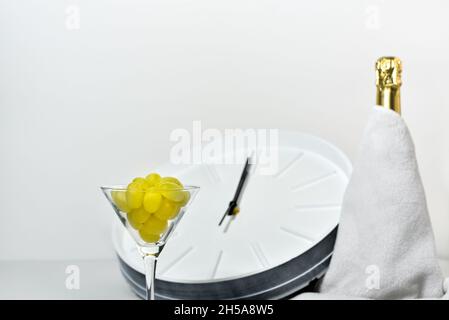 Weinglas mit Trauben auf dem Hintergrund einer verschwommenen Uhr und einer Flasche Champagner. Spanische Tradition, zwölf Trauben zu essen, um Neujahr zu feiern. Stockfoto