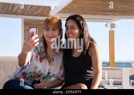 Zwei lateinische Freunde sitzen lächelnd auf einer Terrasse und unterhalten sich bei einem Videoanruf. Stockfoto