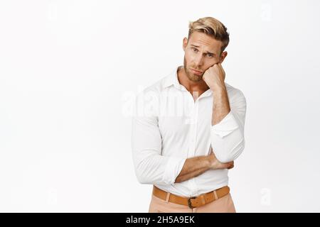 Trauriger und düsterer blonder junger Mann, der vor der Kamera schwül und verzweifelt aussagt, steht in stilvollem weißen Hemd vor Studiohintergrund Stockfoto