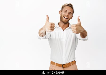 Enthusiastischer Geschäftsmann winkend und lächelnd mit Daumen nach oben, trägt schickes Hemd und Hose, weißer Hintergrund Stockfoto