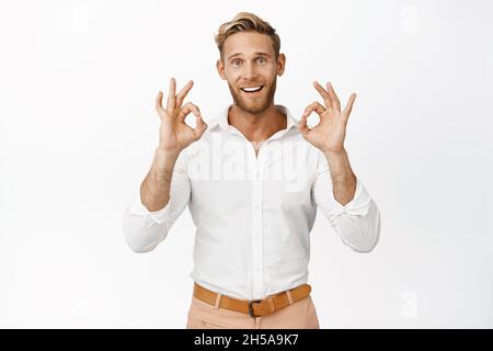 Porträt eines lächelnden blonden Mannes im Hemd, zeigt gute Schilder, geben Zustimmung, zufrieden mit smth, vor weißem Hintergrund stehen Stockfoto