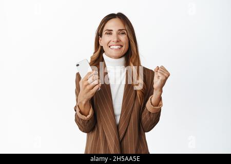 Zufriedene lächelnde Frau mittleren Alters, Geldpreis auf Handy zu gewinnen, Smartphone und Faustpumpe mit fröhlichem Gesicht, weißer Hintergrund halten Stockfoto