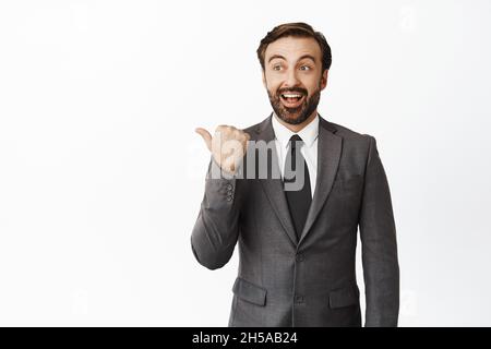 Enthusiastischer Firmenmann in Anzug zeigt und schaut nach links, lächelt erstaunt, prüft Verkauf, Firmenwerbung, weißer Hintergrund Stockfoto