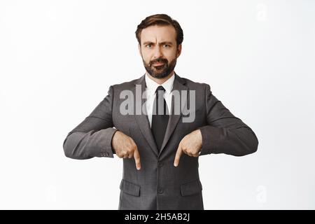 Verwirrt bärtiger Mann in Anzug zeigt die Finger nach unten. Verkäufer zeigt Werbung mit skeptischem Gesichtsausdruck, weißer Hintergrund Stockfoto