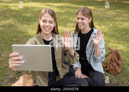 Fröhliche blonde Zwillinge mit Tablet winkende Hände, während jemand während eines Videoanrufs im Park begrüßt Stockfoto