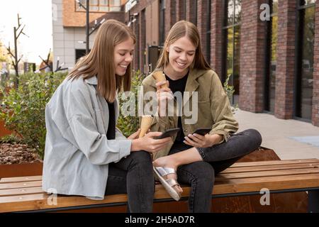 Glückliche Zwillingsschwestern mit Eiscreme-Scrolling auf Smartphones, während sie auf der Bank im städtischen Umfeld sitzen Stockfoto