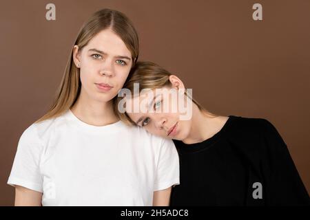Eines der blonden Zwillingsmädchen in weißen und schwarzen T-Shirts, die den Kopf auf der Schulter ihrer Schwester vor braunem Hintergrund halten Stockfoto