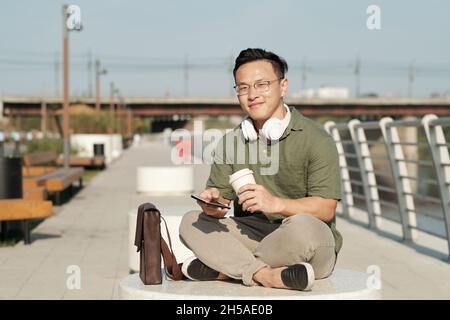 Glücklicher junger Mann in Casualwear, der am heißen Sommertag im Smartphone trank und scrollte, während er sich in der städtischen Umgebung ausruhte Stockfoto